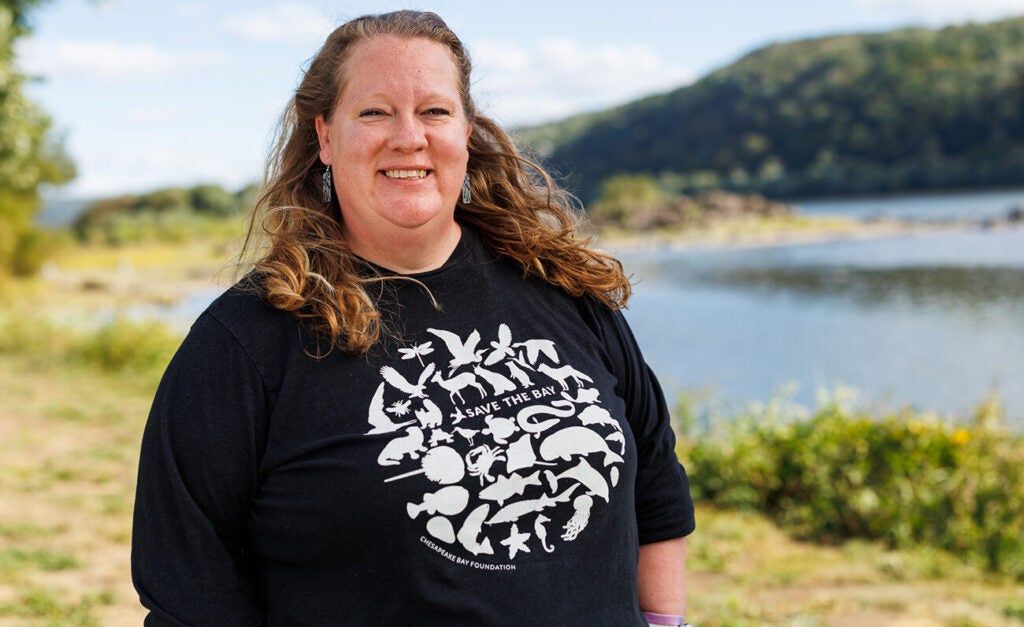 A woman stands by a river, wearing a dark blue sweatshirt with graphics of animals and the words Save the Bay.