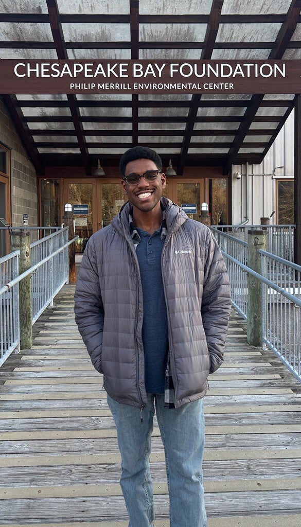 A smiling Black man wearing glasses in a gray Columbia jacket stands on a ramp under a sign reading Chesapeake Bay Foundation.