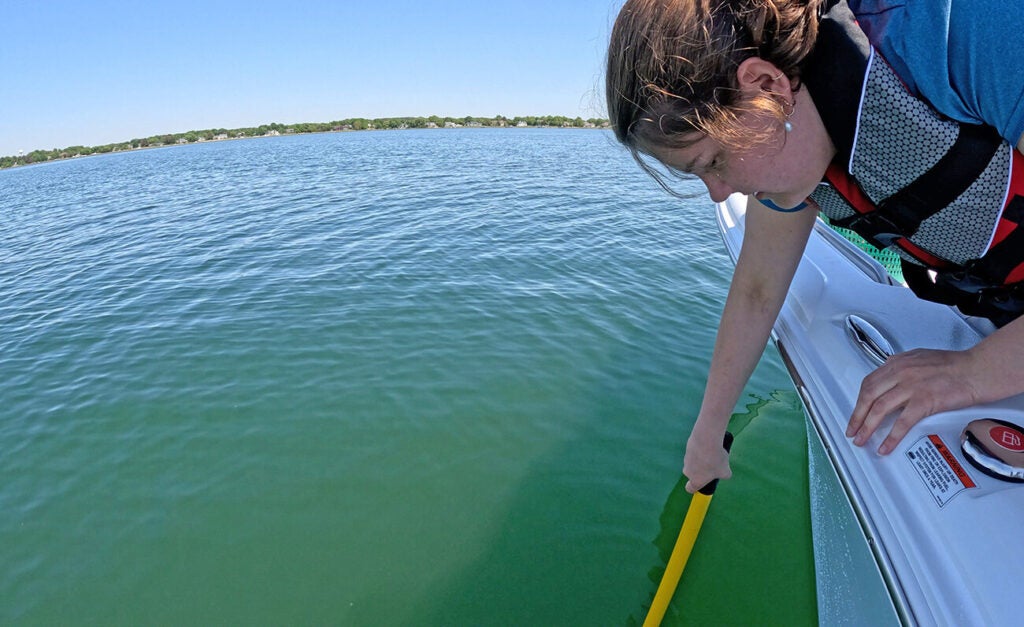 A person wearing a life jacket leans over the side of a boat and dips a yellow pole into the calm green-blue water with a tree-lined shoreline in the distance.