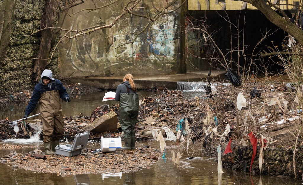 Two people in sweatshirts and waders stand on a small spit of dirt and gravel. One holds a piece of equipment while the other flips through a document. Trash hangs of the bare branches of a bush next to them.
