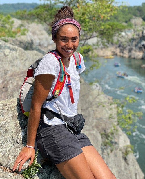 A smiling young woman wearing a colorful headband and backpack sits on a rocky cliff overlooking a river with several rafts in the background.