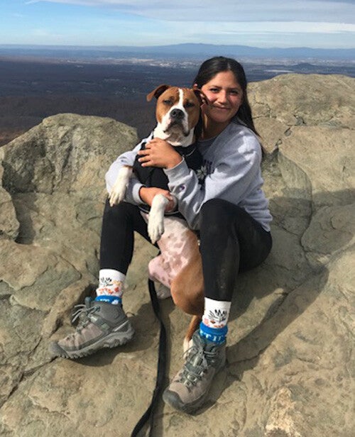 A woman sits on a rocky mountaintop smiling as they hug a brown-and-white dog with a wide valley and cloudy blue sky stretching out behind them.