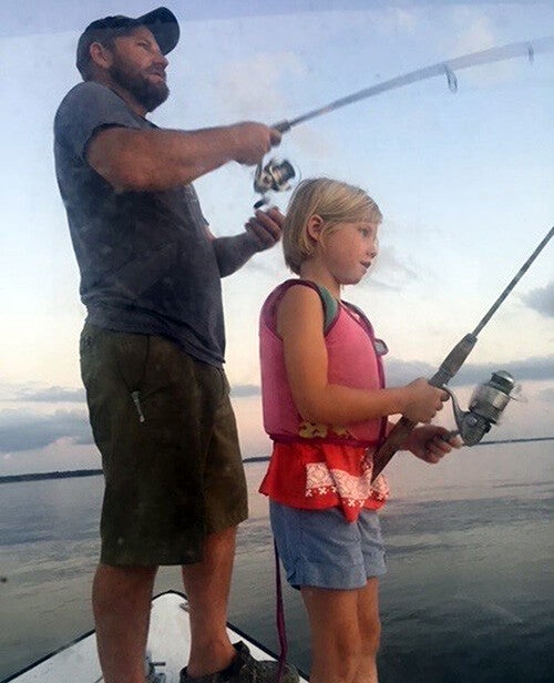 A bearded man and a young girl stand at the bow of a boat, each holding a fishing rod as they fish on calm water at dusk.