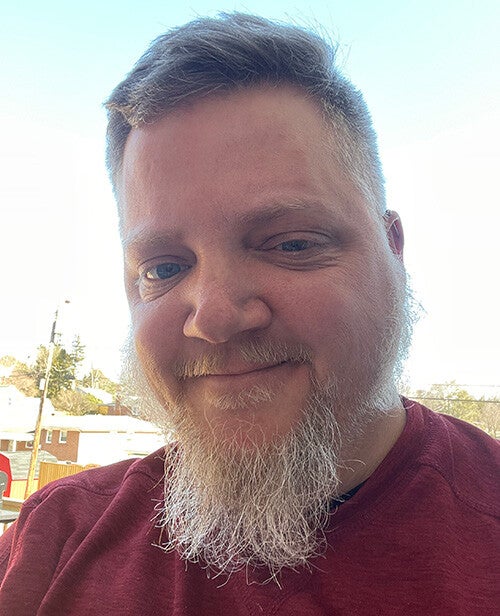 A close-up selfie showing a smiling, light-skinned bearded person wearing a maroon shirt with a short haircut against a bright outdoor background.