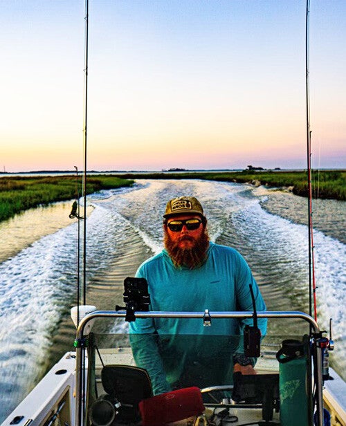 A bearded man wearing a cap and sunglasses steers a small boat through a marshy channel at sunset, leaving twin wakes behind.