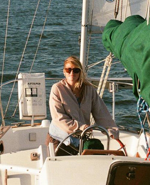 A woman wearing sunglasses steers a sailboat across choppy water on a sunny day, with rigging, a green sail cover, and a distant shoreline visible.