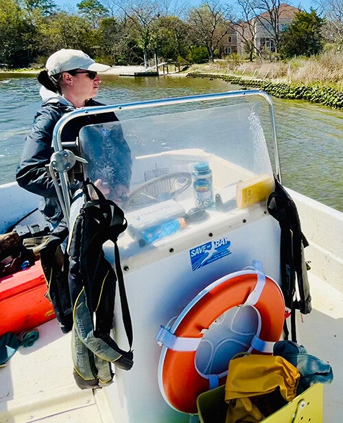 A person wearing a cap and sunglasses steers a small motorboat past a grassy shoreline and houses under a clear blue sky.
