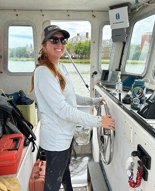 A smiling woman wearing sunglasses and a baseball cap steers a small boat from the cabin, surrounded by navigation equipment, gear, and water visible through the windows.