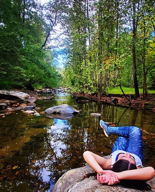 A woman in blue patterned leggings relaxes lying on a large rock beside a calm, tree-lined stream with reflections of the sky and surrounding forest in the water.