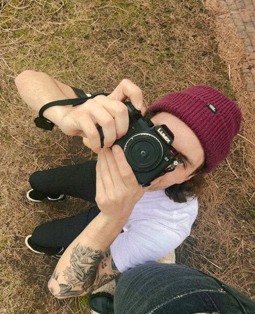 A person wearing a maroon beanie and white shirt kneels on dry grass and points a Lumix camera upward toward the photographer, revealing a tattooed forearm.