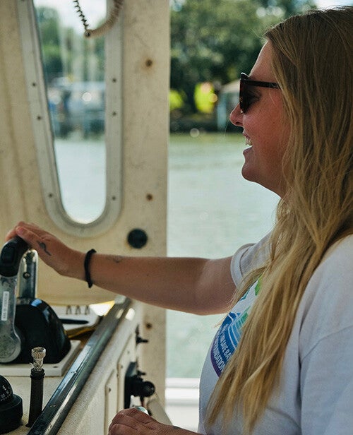 A smiling woman with sunglasses and tattoos steers a boat from the helm, hands on the wheel and throttle with water and shoreline visible through the cabin window.