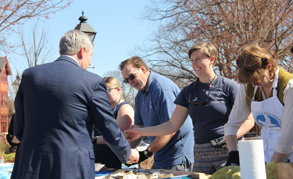 A group of people are outdoors shucking oysters while a smiling woman hands one to a man in a suit.