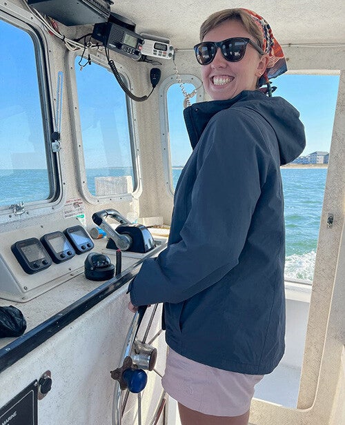 A smiling person wearing sunglasses and a colorful headscarf steers a boat from the cabin while ocean and shoreline are visible through the windows.