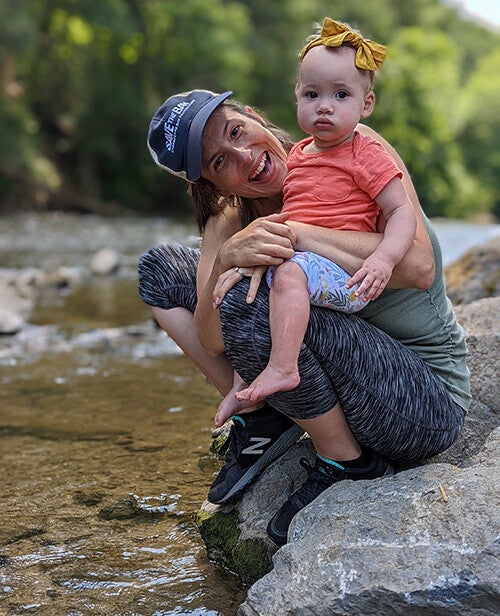 A smiling woman in a baseball cap crouches on a riverside rock while holding a serious-looking baby wearing a yellow bow, with a wooded riverbank in the background.
