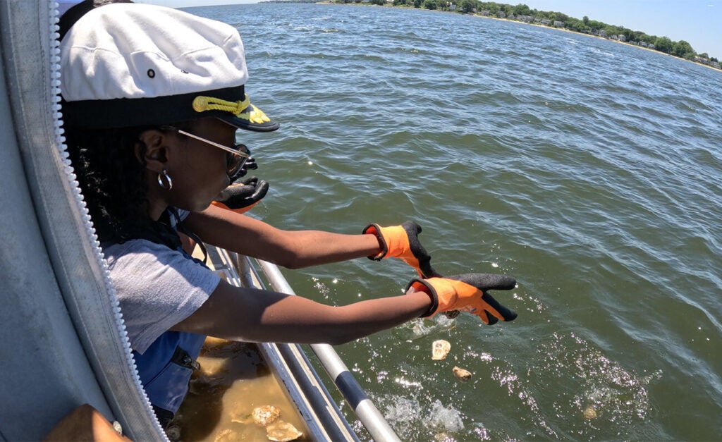 A young Black girl wearing a captain's cap tosses oyster shells over the side of a boat and into the water.