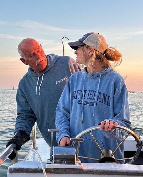 A woman with a blue Smith Island sweatshirt stands at the helm of a small boat, steering, while looking over her shoulder at a man standing behind her.