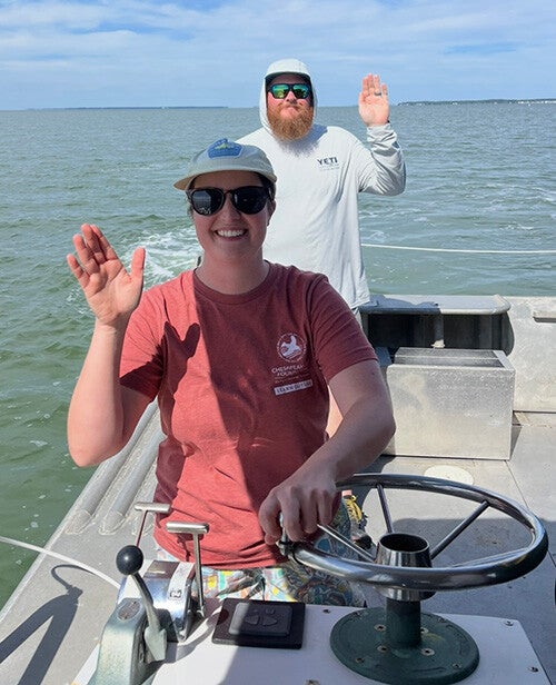 Two smiling people wearing sunglasses and hats wave from the deck of a boat as they steer across calm open water under a blue sky.