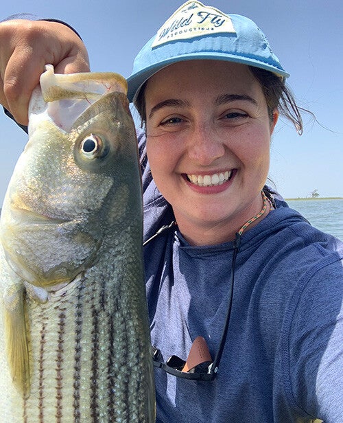 A smiling person in a light-blue cap holds a large striped fish up close to the camera on a sunny day.