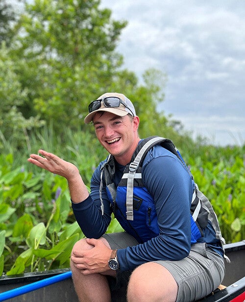 A smiling man wearing a cap, sunglasses, and a life jacket sits in a canoe surrounded by lush marsh plants under a cloudy sky.