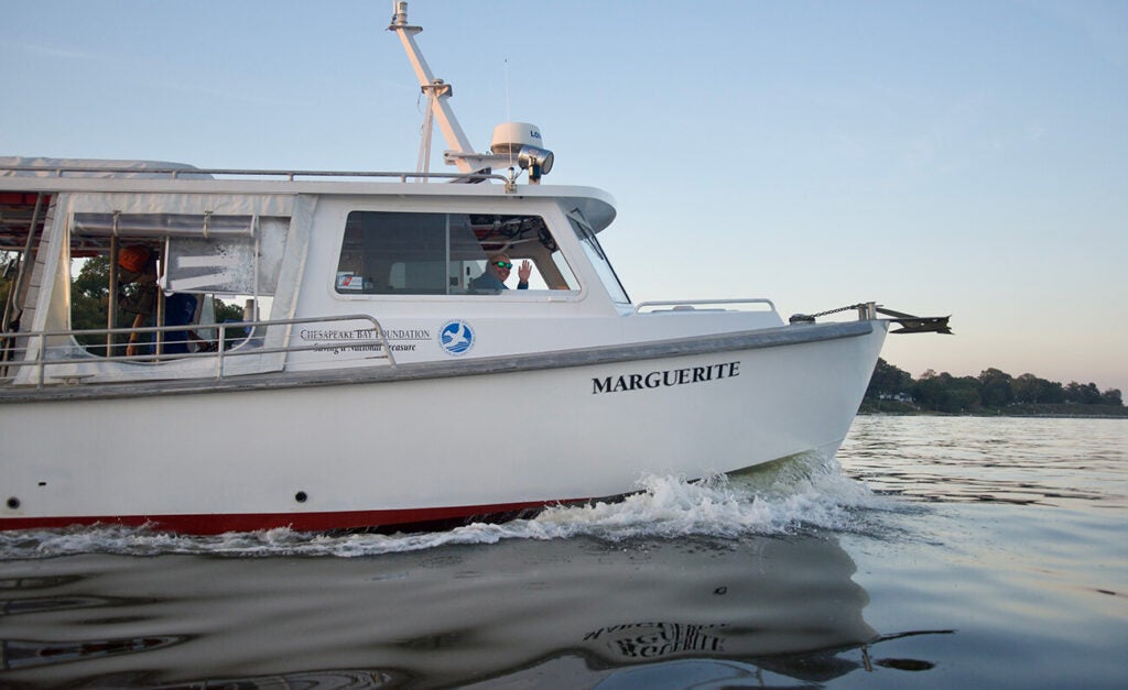 A white boat named “MARGUERITE” with a Chesapeake Bay Foundation logo cruises on calm water while a person wearing sunglasses waves from the cabin.