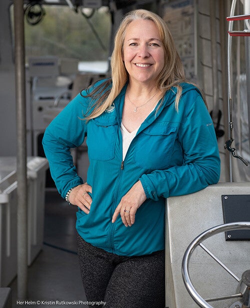 A smiling woman in a teal jacket and anchor necklace stands with her hand on her hip near the helm aboard a boat.