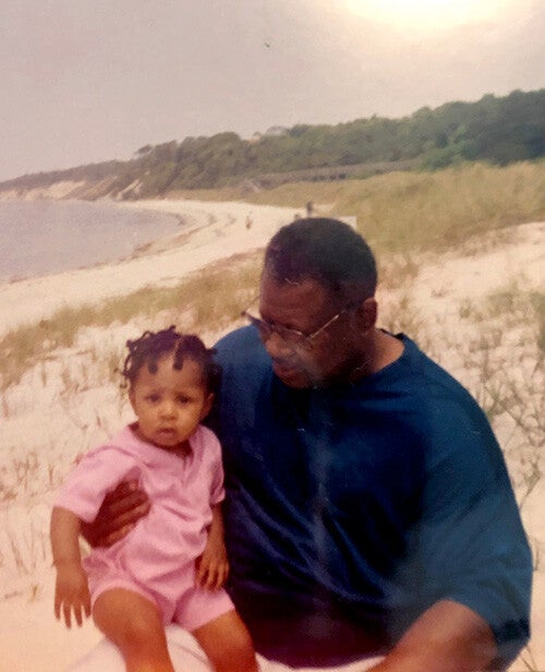 A man holding a young child while sitting on a sandy beach with grassy dunes and a bright sun in the sky.