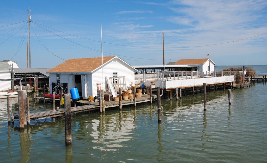 A row of white wooden shacks and docks sits over calm water under a bright blue sky.