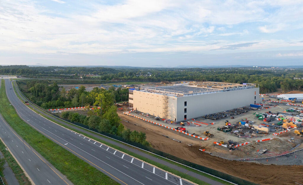 A large industrial building under construction sits beside a multilane highway, surrounded by construction equipment, orange barriers, and a wide, partly cloudy sky.
