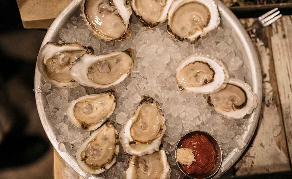 Eleven shucked oysters sit in a bowl of ice along with a cup of cocktail sauce.
