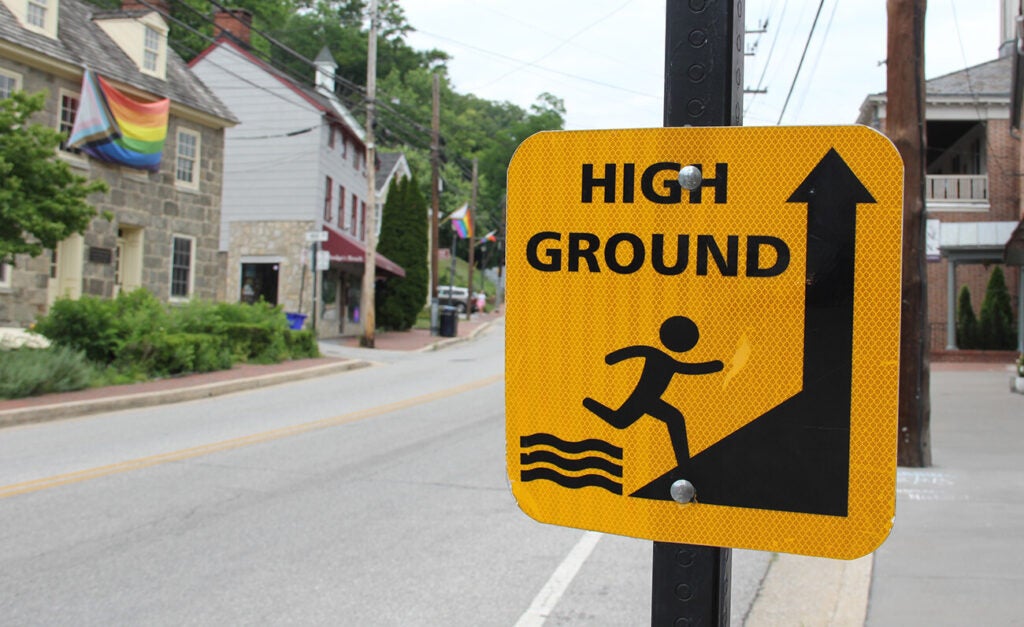 A close-up of a yellow “HIGH GROUND” warning sign mounted on a pole with a small-town street, buildings, and a rainbow flag visible in the background.