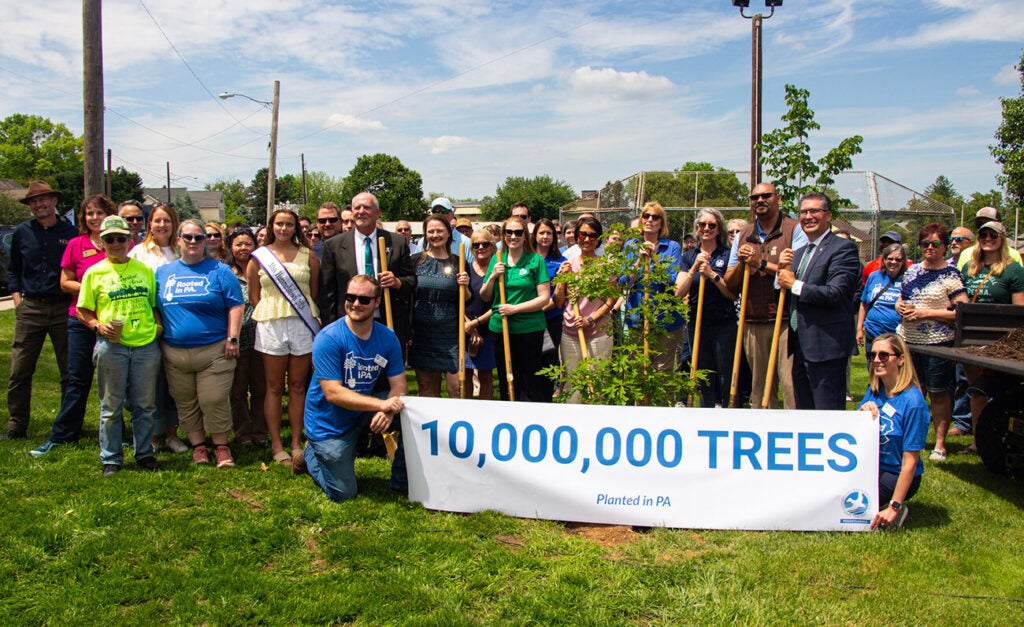 A crowd of people stand behind a tree sapling as two people kneel in front of it, holding a banner that reads 10,000,000 Trees Planted in PA.