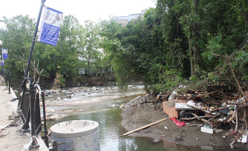 A river flows along a concrete walkway with debris piled up around it.
