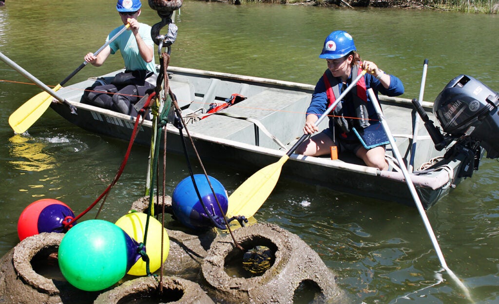 Two people wearing blue hard hats sitting in a dinghy use paddles to maneuver four concrete reef balls with colorful buoys into place on a calm river.