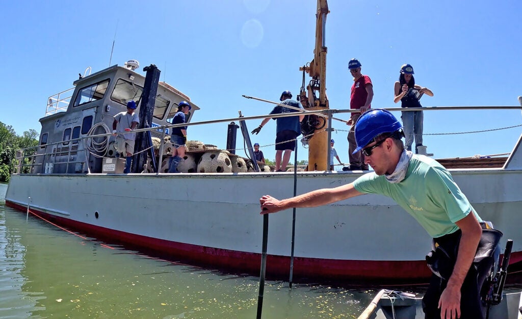 A crew of people wearing hard hats work on a workboat installing concrete reef modules while a man in the foreground leans over the water holding a pole.