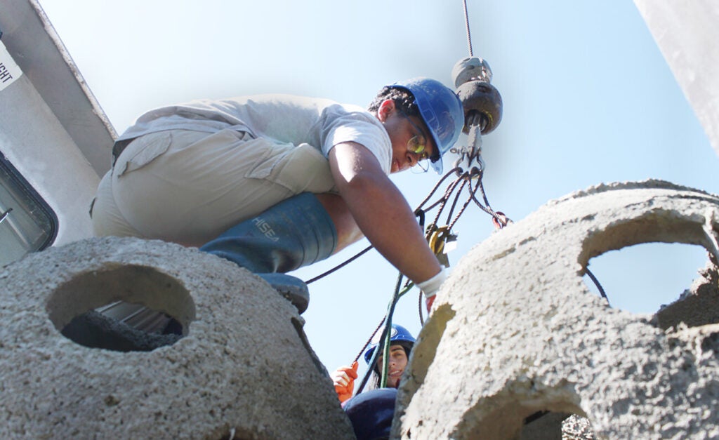 A man in a blue hard hat crouches atop a concrete reef ball attaching cables, while a coworker can be seen in the background.