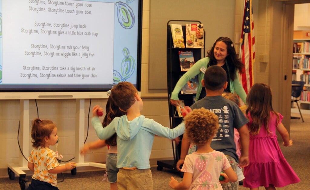 Young children actively participate in an interactive library storytime, dancing along to an ocean-themed rhyme displayed on screen while the storyteller leads the fun.
