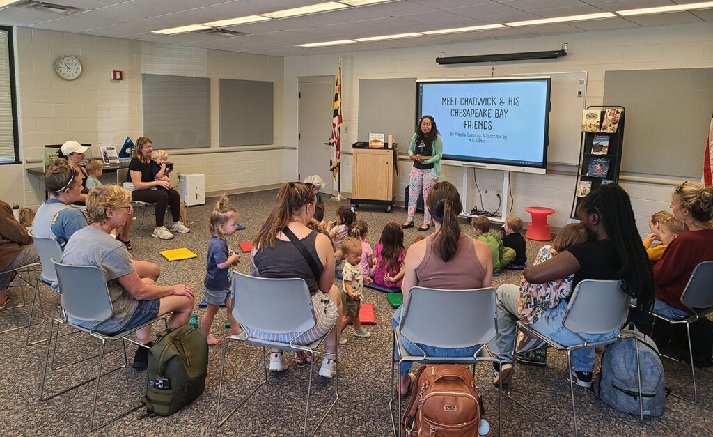A presenter leads an interactive "Meet Chadwick & His Chesapeake Bay Friends" storytime session for a group of young children and their parents seated in a circle.