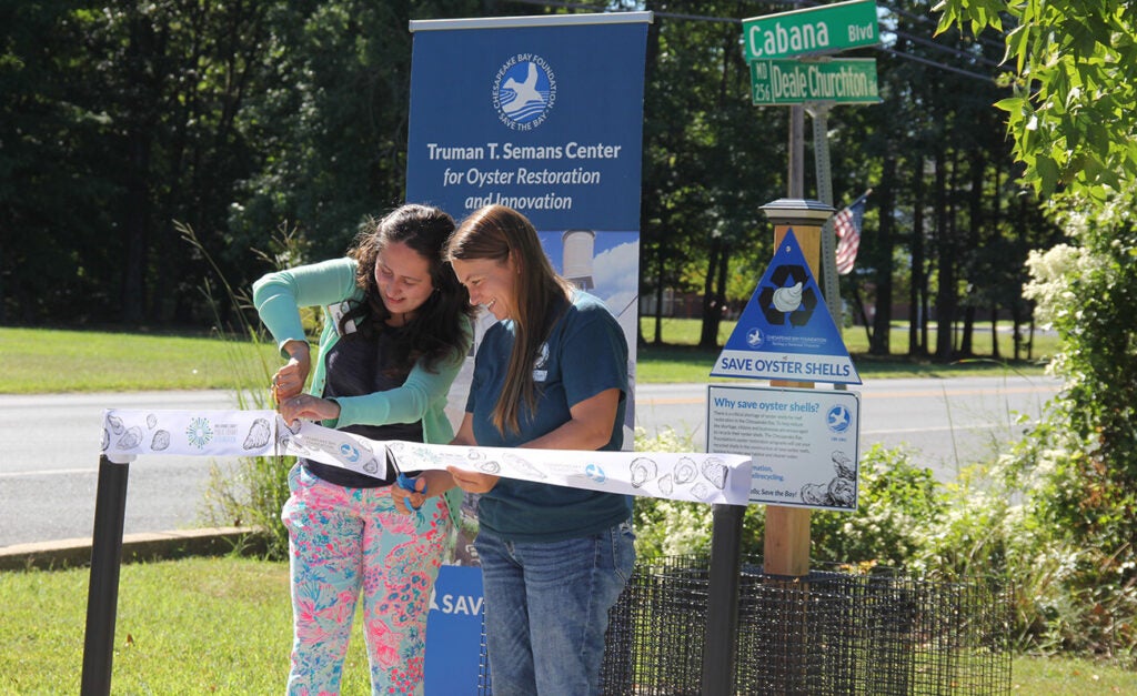 Two smiling women cut the ceremonial ribbon to celebrate the opening of a new Chesapeake Bay Foundation oyster shell recycling station.