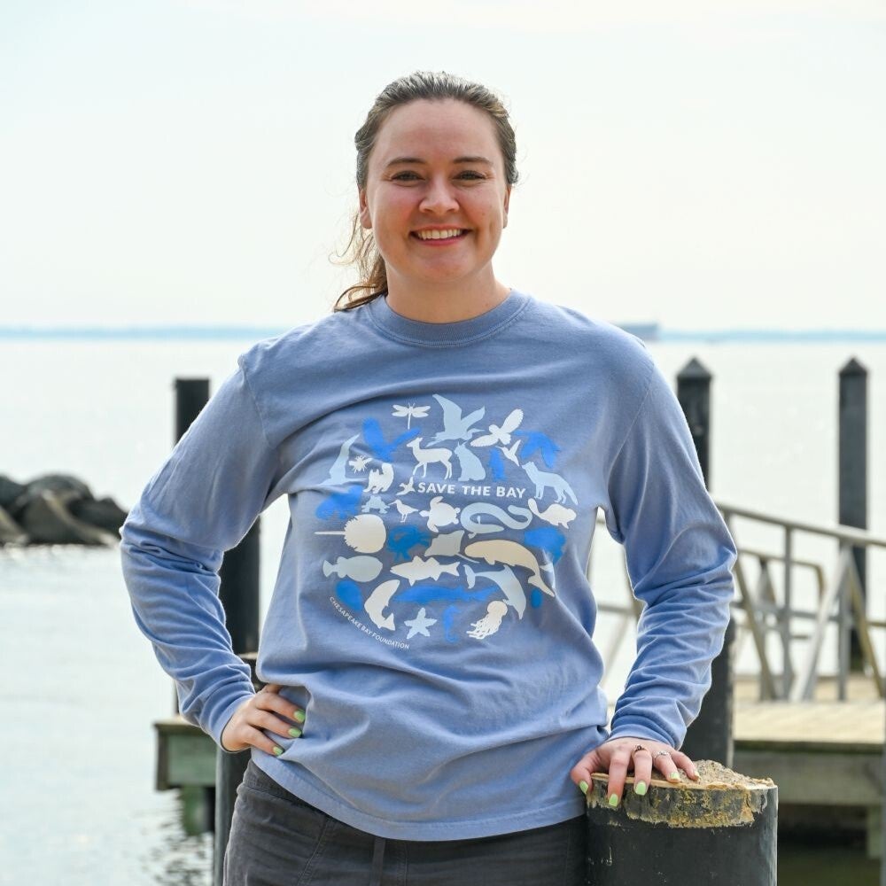 A woman stands at a dock wearing a blue t-shirt with images of animals and fish and the words Save the Bay.