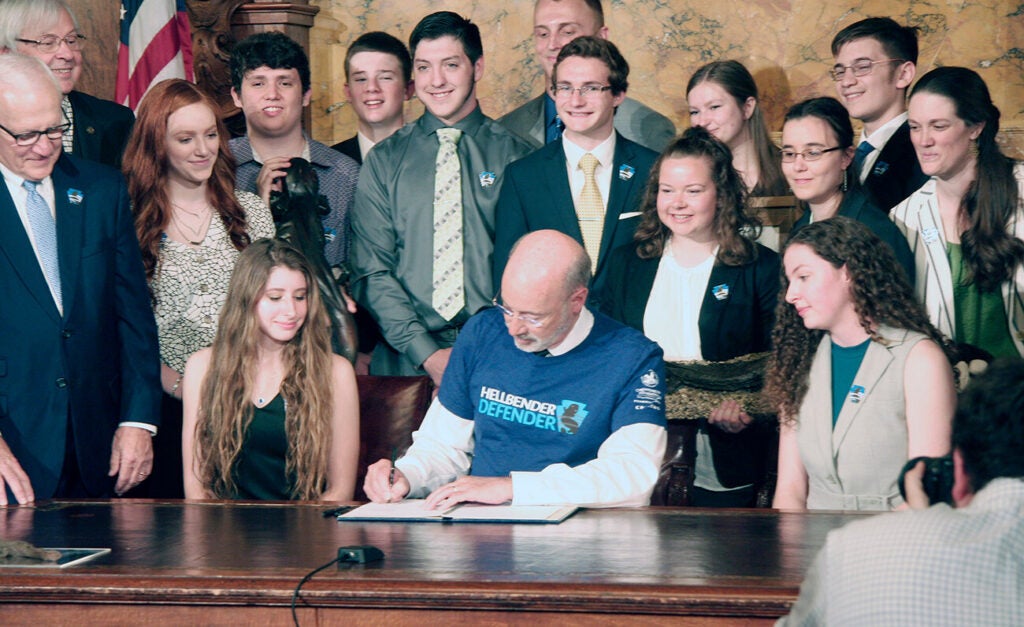 Former Governor Tom Wolf wears a blue Hellbender Defender shirt and signs legislation surrounded by young advocates at a bill signing ceremony.