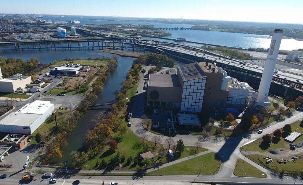 Aerial view of an industrial facility near a waterway, with surrounding roads, bridges, and buildings; a large smokestack and green spaces are visible, and a wide river extends toward the horizon under clear skies.
