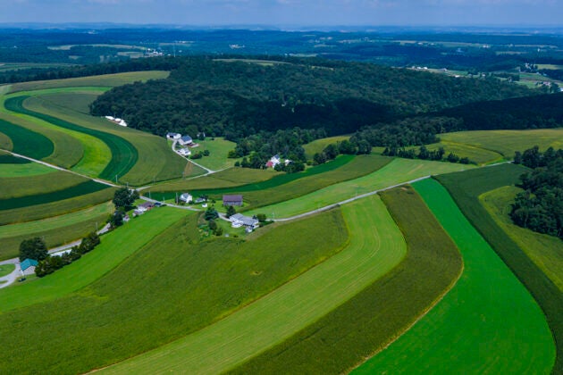 An aerial view shows contoured farm fields in varying shades of green rolling across the landscape with forests and farmhouses interspersed throughout the land