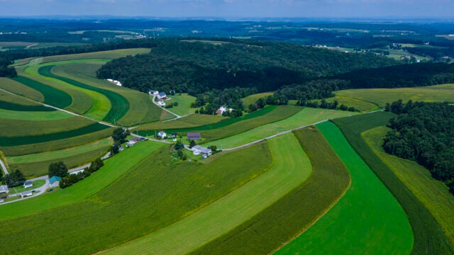 An aerial view shows contoured farm fields in varying shades of green rolling across the landscape with forests and farmhouses interspersed throughout the land.