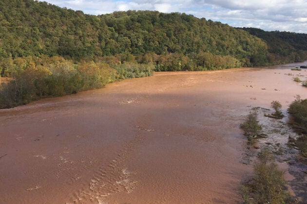 The Susquehanna River overflows its banks with muddy brown floodwaters submerging trees and vegetation along the forested shoreline.