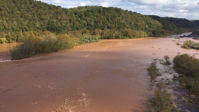 The Susquehanna River overflows its banks with muddy brown floodwaters submerging trees and vegetation along the forested shoreline.