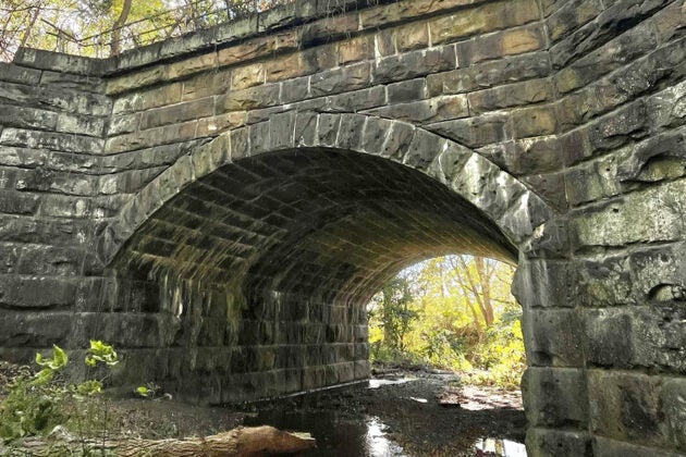 Weathered stone arch bridge over a stream with moss-covered stonework and sunlight filtering through trees beyond.