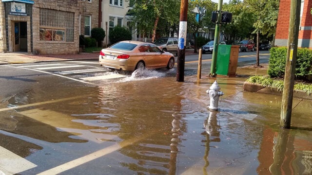 A car drives through floodwater on a street past a fire hydrant partially submerged in the water.