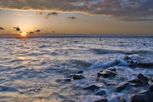 Sunset over a body of water with waves breaking over rocks and golden light streaming through clouds on the horizon.