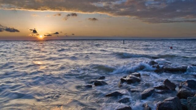 Sunset over a body of water with waves breaking over rocks and golden light streaming through clouds on the horizon.