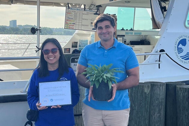 Two people pose in front of a boat, one holding a certificate and the other holding a potted plant, with the Chesapeake Bay Foundation logo visible in the background.